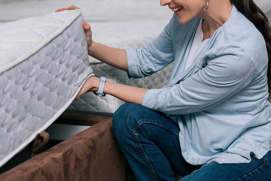 A dark-haired woman wearing a light blue cardigan and jeans lifts the corner of a mattress.